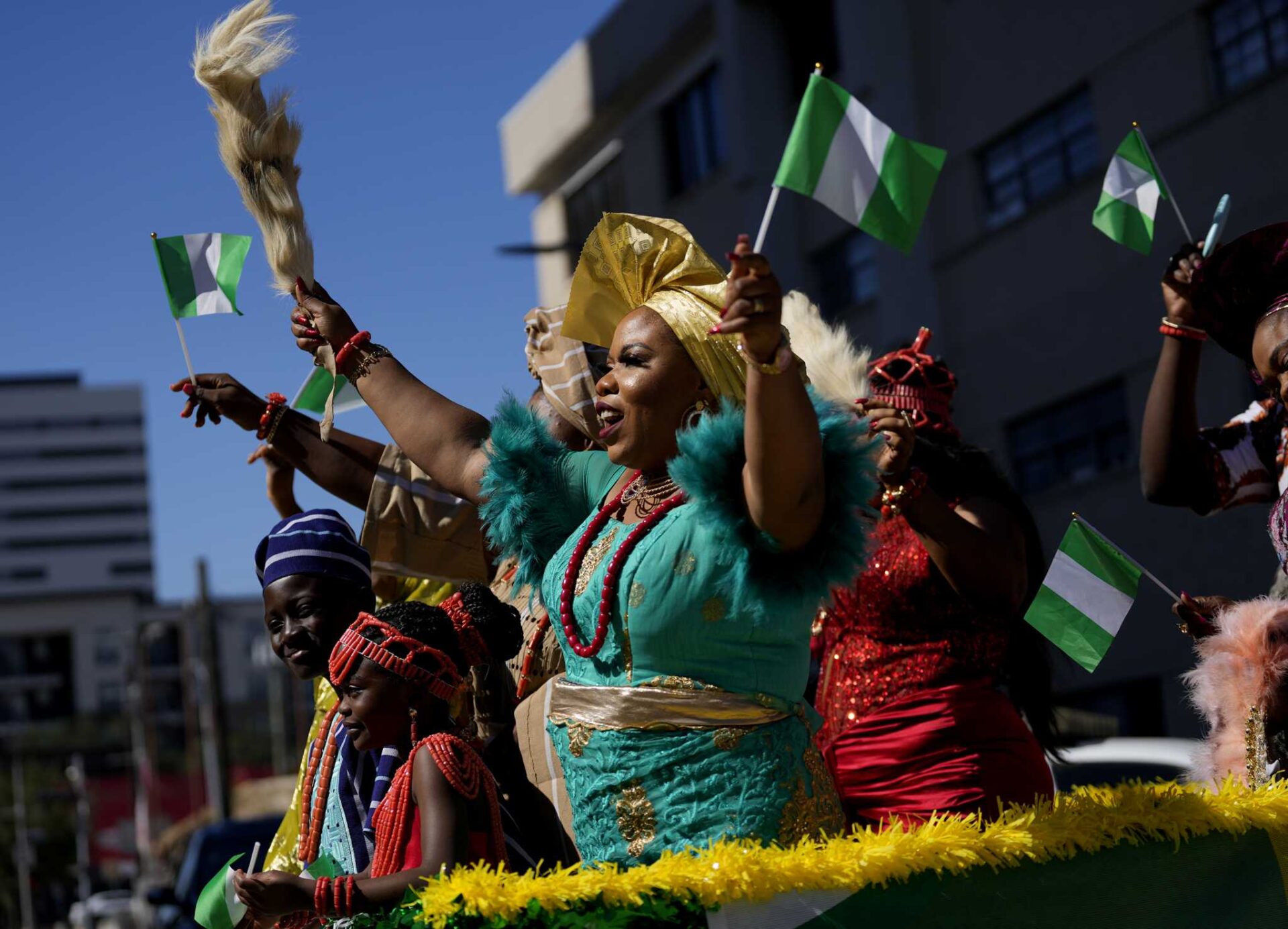 A woman in turquoise and gold attire waves a whisk as celebrants in traditional dress raise Nigerian flags against a blue sky.