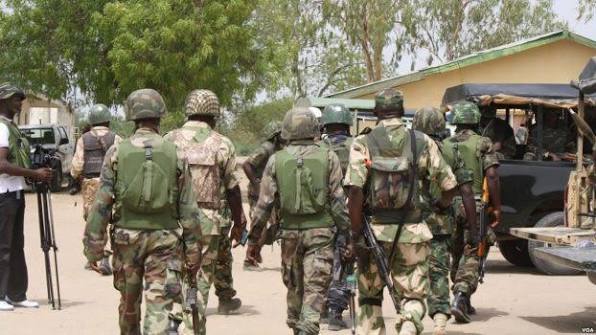 Nigerian troops in camouflage and helmets walk together beside military vehicles near a low building, with trees in the background.